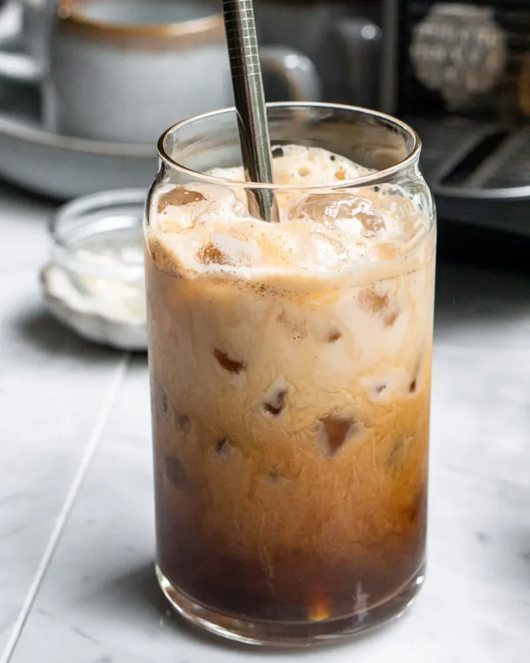 A tall glass of iced French toast latte with a metal straw sticking out of it. there is a Nespresso machine behind the glass.