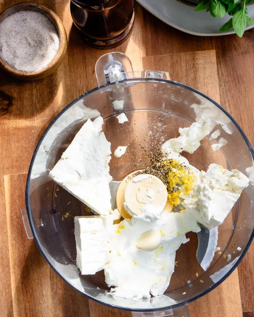 A food processor with a block of feta cheese, lemon zest, salt, black pepper ready to be blended.