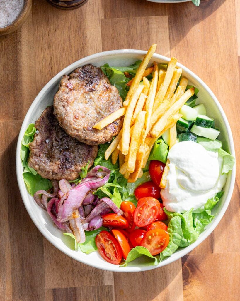 A mediterranean lamb bowl with lamb burgers, french fries, whipped feta, cherry tomatoes.