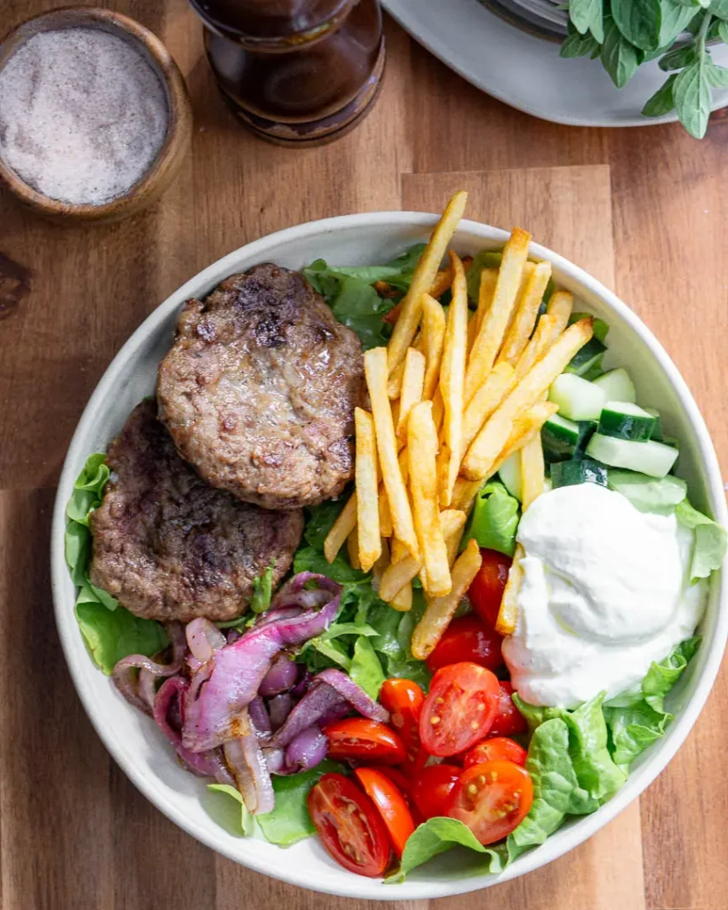A shallow bowl filled with lettuce and topped with 2 lamb burgers, grilled red onions, cherry tomatoes, cucumbers, whipped feta, and french fries.