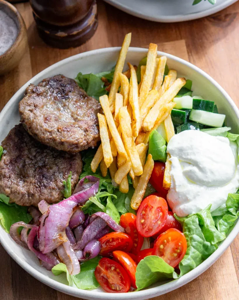 A lamb bowl with all the toppings including, lamb burgers, whipped garlic feta, french fries, onions, tomatoes and cucumbers.