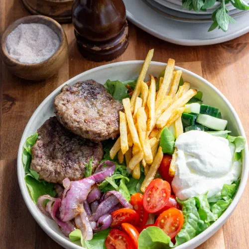 A bowl of lettuce topped with all the ingredients to make a Mediterranean lamb bowl. Lamb burgers, veggies, french fries, and whipped feta.