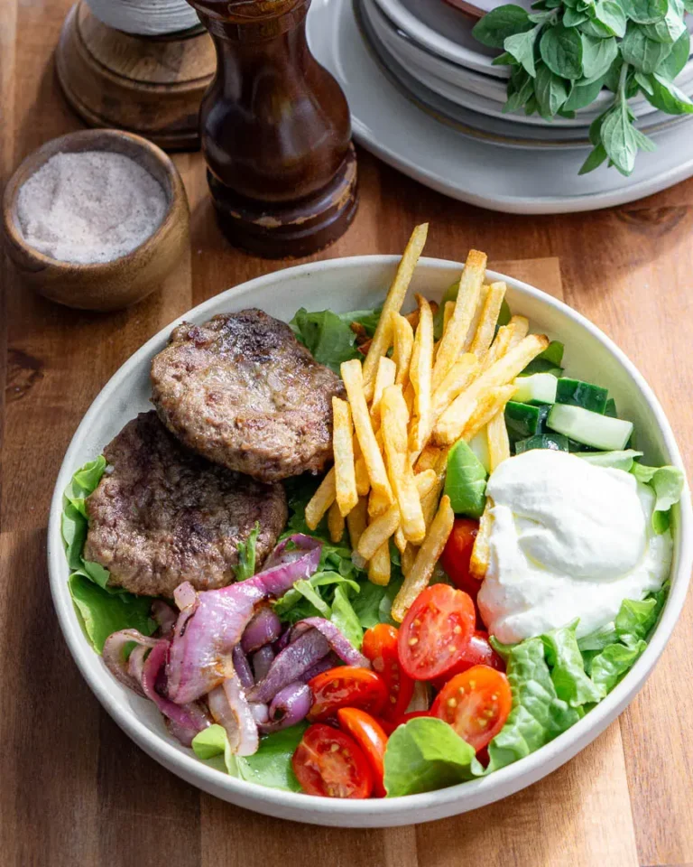 A bowl of lettuce topped with all the ingredients to make a Mediterranean lamb bowl. Lamb burgers, veggies, french fries, and whipped feta.