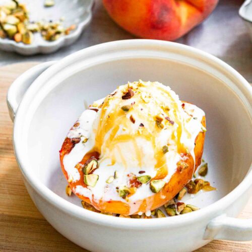 A grilled peach in a bowl topped with a scoop of vanilla ice cream, honey drizzle, and chopped pistachios.