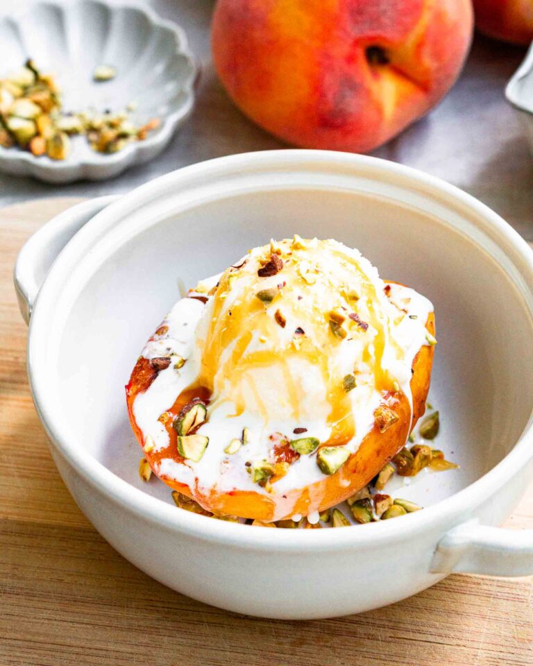 A grilled peach in a bowl topped with a scoop of vanilla ice cream, honey drizzle, and chopped pistachios.