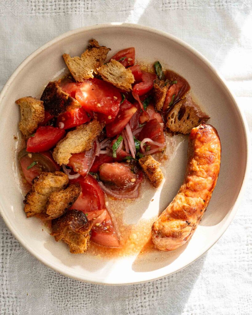 A plate with a simple tomato salad with sourdough croutons. There is a grilled Italian Sausage link next to the salad.