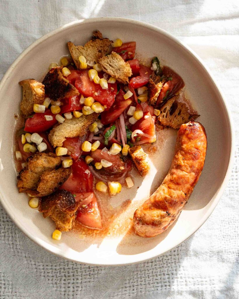 Simple tomato salad topped with corn and croutons on a plate next to Italian sausage.