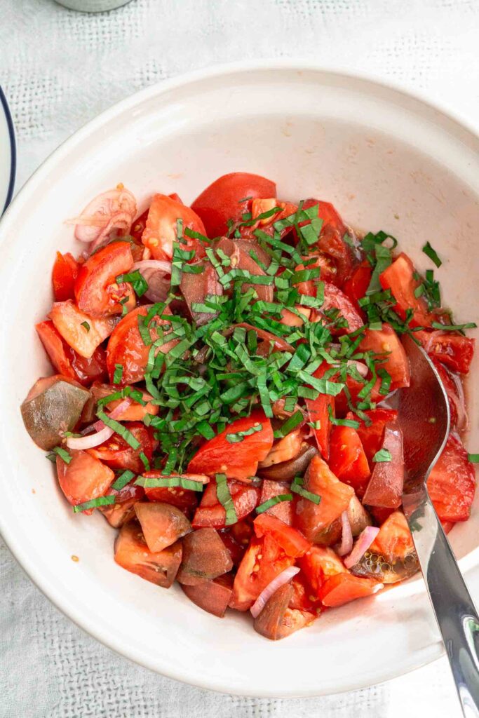 Basil chiffonade on top of large chunks of tomatoes that are in a bowl. There is a stainless steel spoon in the bowl for serving.