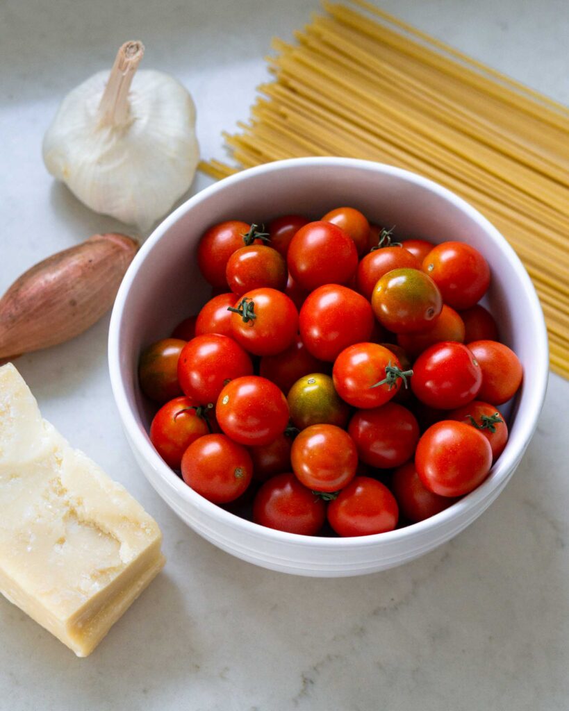 A bowl of cherry tomatoes surrounded by spaghetti, one shallot, a garlic bulb, and a wedge of parmesan cheese.
