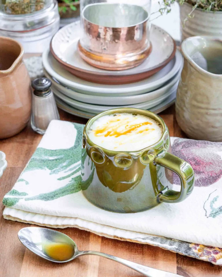 A warm mug of salted honey latte on a tea towel. There is a spoon laying in front of the mug and it has a bit of honey on it.