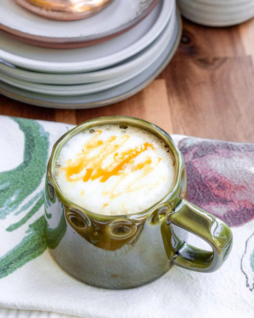 A hot mug of salted honey latte with a caramel drizzle. The mug is sitting on a dish towel.
